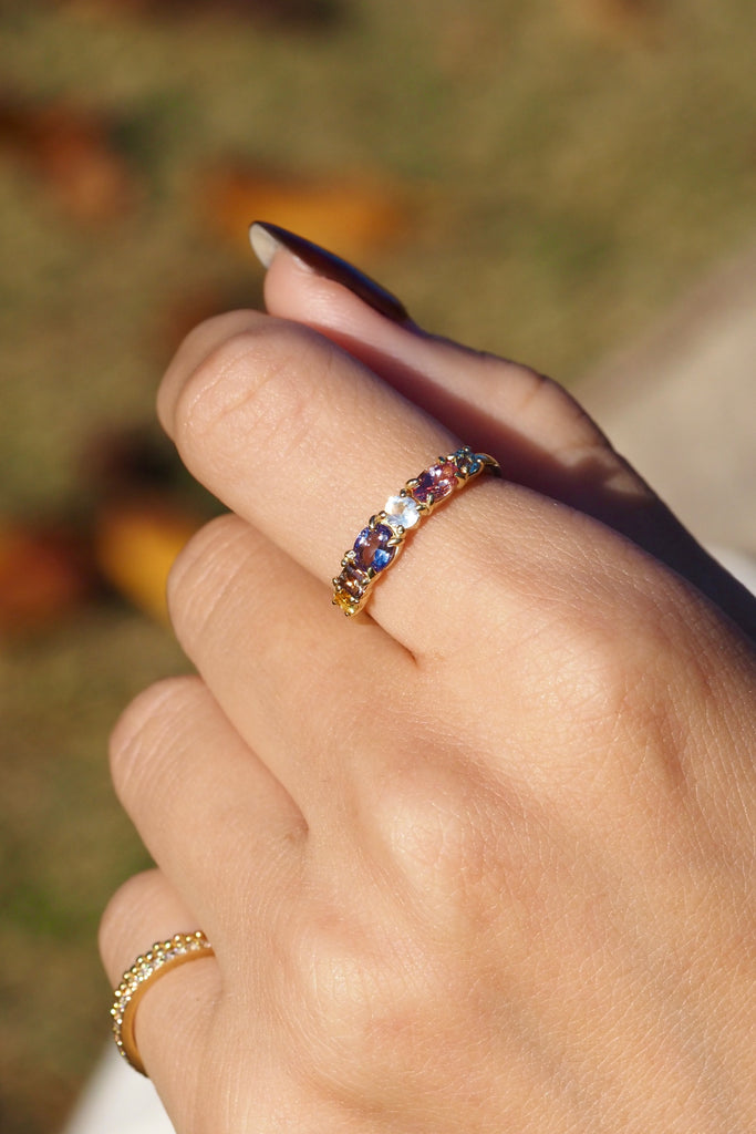 ジュエリースタジオMASHIROのアップサイクルジュエリー。マラヤガーネット・オーバル・約0.3ct・約 5*3mm・タンザニア産｜タンザナイト・オーバルカット・約0.28ct・約4.9*3.8mm｜レインボーームーンストーン・ラウンド・約0.09ct・約3mm｜マルチカラーサファイア（３石：ラウンド・プリンセス・ヘキサゴン）・合計：0.34ct・約2.6mm。シャンパンゴールドカラーのカクテルエタニティリングの着用画像。