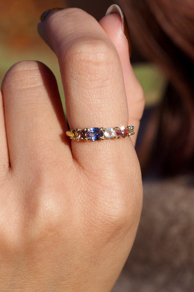 ジュエリースタジオMASHIROのアップサイクルジュエリー。マラヤガーネット・オーバル・約0.3ct・約 5*3mm・タンザニア産｜タンザナイト・オーバルカット・約0.28ct・約4.9*3.8mm｜レインボーームーンストーン・ラウンド・約0.09ct・約3mm｜マルチカラーサファイア（３石：ラウンド・プリンセス・ヘキサゴン）・合計：0.34ct・約2.6mm。シャンパンゴールドカラーのカクテルエタニティリングの着用画像。